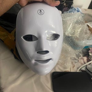 K&ouml;nnte beinhalten: Eine wei&szlig;e LED-Gesichtsmaske mit &Ouml;ffnungen f&uuml;r Augen, Nase und Mund. Die Maske hat einen ber&uuml;hrungsempfindlichen Ein-/Ausschalter. Ein schwarzes Smartphone und andere Gegenst&auml;nde befinden sich im Hintergrund.