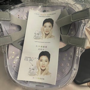 K&ouml;nnte beinhalten: Eine transparente Lichttherapie-Gesichtsmaske mit grauen, verstellbaren Tr&auml;gern. Die Maske hat zahlreiche kleine LED-Leuchten und enth&auml;lt ein gedrucktes Anleitungsblatt mit Text und Illustrationen. Der Text auf dem Blatt lautet "LIGHT BEAUTY INSTRUMENT".