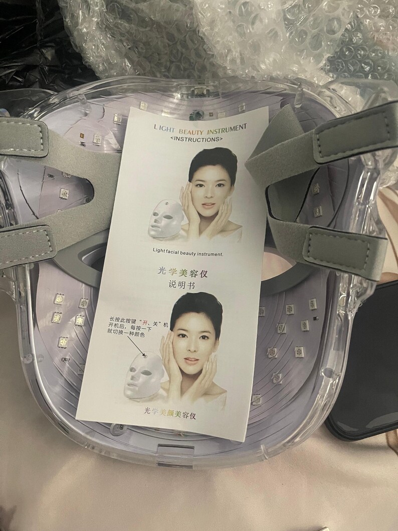 K&ouml;nnte beinhalten: Eine transparente Lichttherapie-Gesichtsmaske mit grauen, verstellbaren Tr&auml;gern. Die Maske hat zahlreiche kleine LED-Leuchten und enth&auml;lt ein gedrucktes Anleitungsblatt mit Text und Illustrationen. Der Text auf dem Blatt lautet "LIGHT BEAUTY INSTRUMENT".