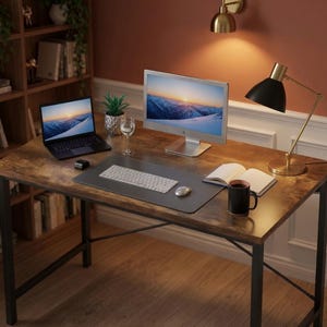May include: A rectangular desk with a dark wood grain surface and black metal frame. The desk holds a laptop, a computer monitor, a white keyboard, a computer mouse, a small potted plant, a glass, an open notebook, and a black mug.