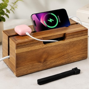 May include: A rectangular wooden cable management box with a natural wood grain finish. The top features a slot holding a smartphone displaying a green charging icon and a pink wireless earbud case connected by a white charging cable to the interior.