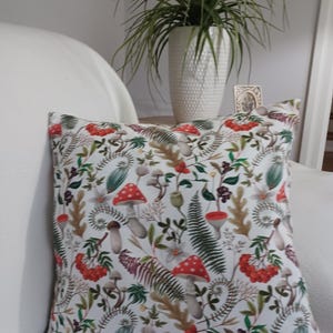 May include: A square scatter cushion featuring a white background with a repeating pattern of red and white spotted mushrooms, green ferns, brown oak leaves, and clusters of small red berries. The design covers the entire front surface of the cushion.