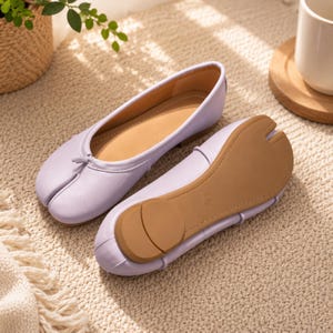May include: A pair of light purple ballet flats featuring a split-toe design, a small decorative bow at the vamp, and a tan sole. The shoes have a smooth finish, a low-profile silhouette, and a visible size marking of 240 on the bottom of the sole.