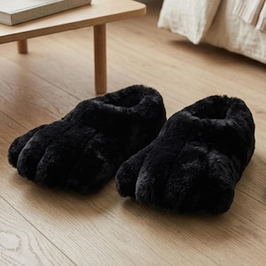May include: A pair of black slippers crafted from plush, faux fur material. The slippers feature a distinct design with padded, rounded toe sections that resemble animal paws, providing a soft, textured appearance and a slip-on construction.