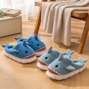 May include: Two pairs of blue shark-shaped slippers with white soles and red and white mouths. The slippers have a soft, plush texture and are designed to resemble sharks. The slippers are on a wooden floor.