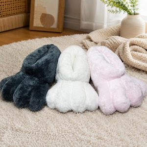 May include: Three plush paw-shaped slippers in a row. The slippers are in three colors: dark gray, white, and light pink. The slippers are made of a soft, fuzzy material and are designed to cover the feet.
