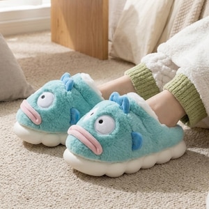 以下が含まれることがあります： 魚の形をしたスリッパ。水色で、白、ピンク、青のアクセントがあります。ふわふわした質感で、大きな目と笑顔の口が特徴的な漫画のデザインです。
