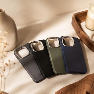 Peut inclure: Quatre coques de smartphone rectangulaires disposées en rangée, présentant une finition mate lisse dans des tons gris foncé, noir, vert olive et bleu marine. Chaque coque comprend un cadre carré métallique argenté pour l'appareil photo.