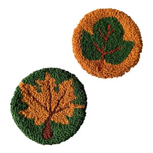 May include: Two circular punch needle coasters featuring textured leaf designs. One coaster displays a golden-orange maple leaf on a dark green background, while the other shows a dark green leaf with orange veins on a golden-orange background.