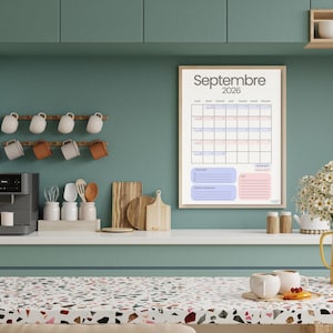 Puede incluir: Un calendario enmarcado de septiembre de 2026 cuelga de una pared turquesa en una cocina. El calendario tiene un fondo beige con una cuadrícula de fechas en azul y rosa. Una jarra amarilla con flores blancas está en la encimera.