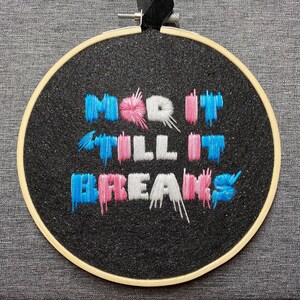 Puede incluir: Arte de bordado en aro con el texto "Mod it till it breaks" en hilo azul, rosa y blanco sobre un fondo negro brillante.