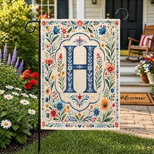 May include: A garden flag with a large blue letter "H" in a decorative frame, surrounded by colourful floral and leaf patterns. The flag is displayed outdoors on a black metal stand. The background shows a house and garden.