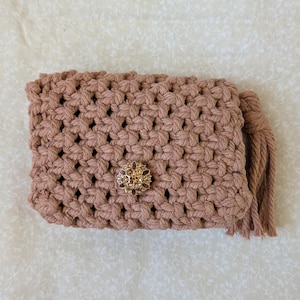 May include: A handmade, light brown macrame clutch with a decorative gold-colored button clasp. The clutch features a woven pattern and a tassel on the side. The bag is rectangular and appears to be made of a soft, natural fiber.