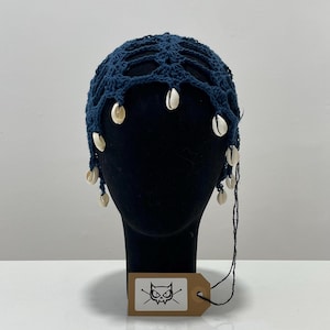Peut inclure: Un bonnet en crochet bleu foncé avec un motif en filet ajouré et des coquillages cauris blancs suspendus le long du bord inférieur. Une étiquette rectangulaire marron avec un graphique noir représentant une tête de chat est attachée.