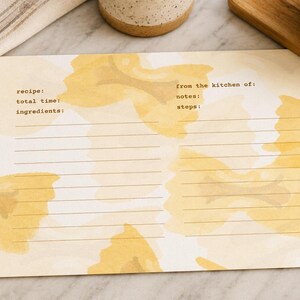 Pode incluir: Um cartão de receita com um design em aquarela em tons de amarelo e creme. O cartão inclui instruções para a receita, tempo total, ingredientes, notas e etapas. Uma colher de pau e um pano de cozinha listrado são visíveis.