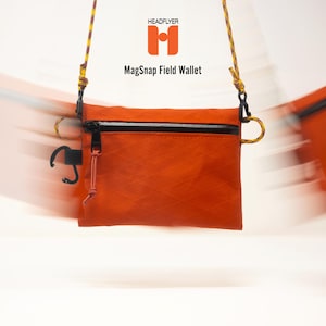 May include: An orange MagSnap Field Wallet by HEADFLYER featuring a diamond-patterned texture, a black zippered top closure, and a black plastic clip on the side. The wallet is suspended by a yellow and orange cord strap against a white background.