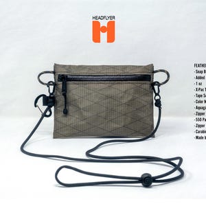May include: A rectangular olive green wallet with a diamond grid pattern, featuring a black zippered top, 550 paracord loops, and a long black cord strap. Text includes HEADFLYER and FEATHER FIELD WALLET with a list of features like 1 oz and X-Pac.