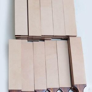 May include: A collection of rectangular leather strops featuring smooth, light beige leather surfaces mounted on dark brown stained wooden handles. Each handle includes a small hole at the base with a looped tan cord for hanging storage.