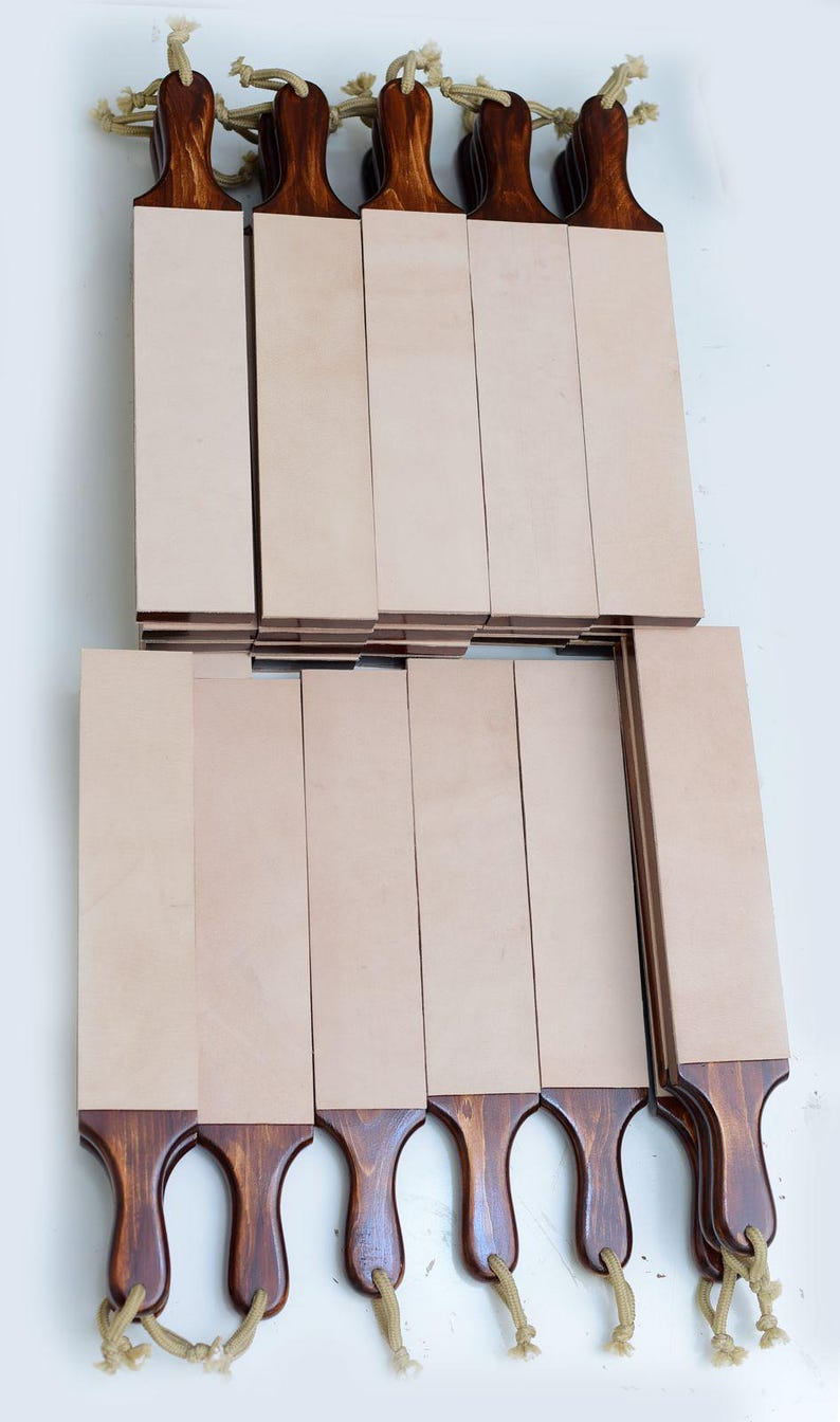 May include: A collection of rectangular leather strops featuring smooth, light beige leather surfaces mounted on dark brown stained wooden handles. Each handle includes a small hole at the base with a looped tan cord for hanging storage.