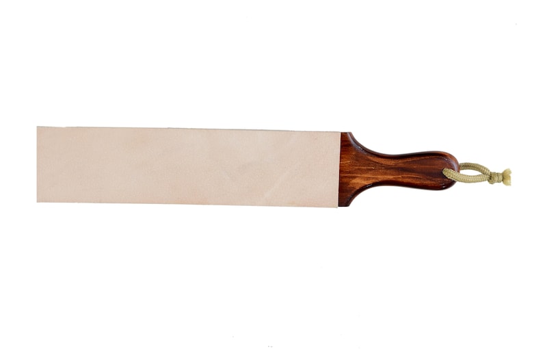 May include: A leather sharpening strop featuring a rectangular light beige leather surface attached to a dark brown polished wooden handle. A short, light-coloured cord loop is threaded through a hole at the end of the wooden handle for hanging.