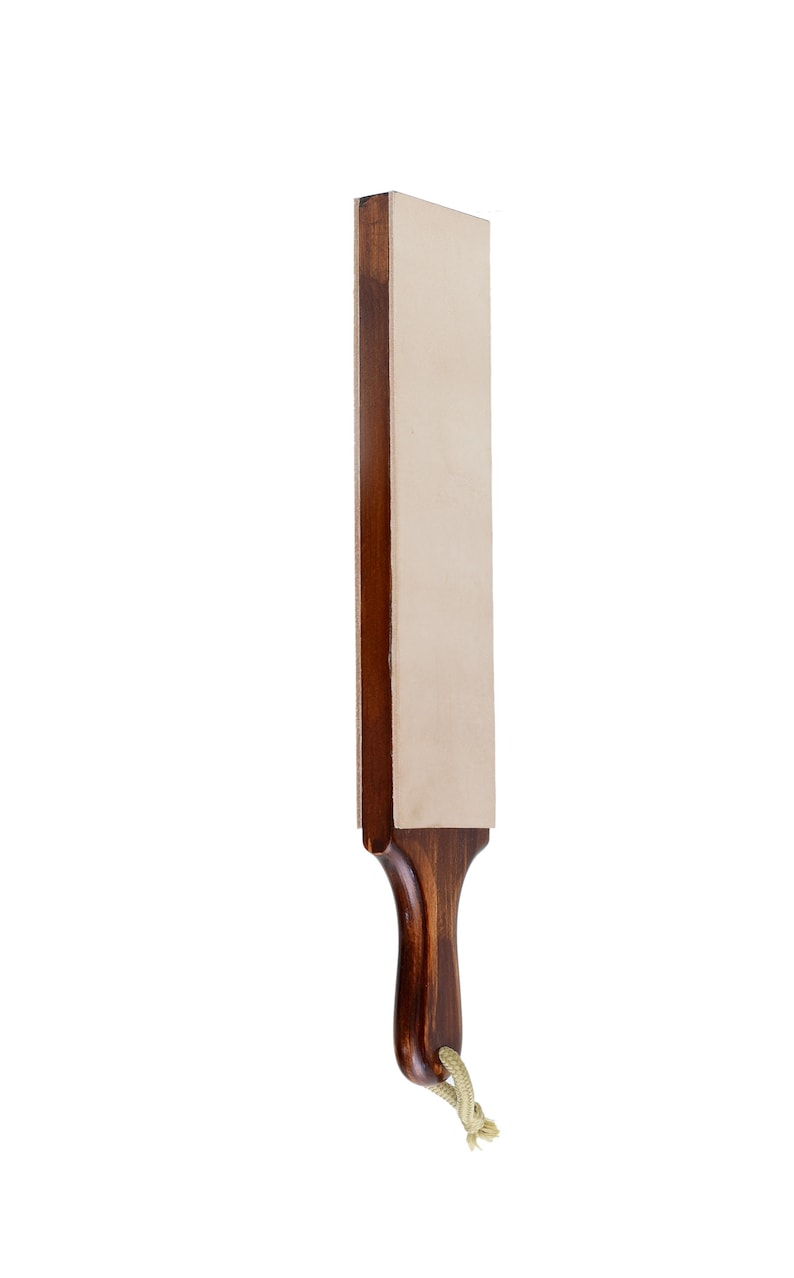 May include: A rectangular leather honing strop featuring a smooth, light beige leather surface mounted on a dark brown wooden handle. The handle has a curved, ergonomic shape and includes a small, light-coloured rope loop at the base for hanging.