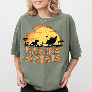 May include: An olive green t-shirt featuring a graphic of a yellow sun setting behind silhouettes of a lion cub, a meerkat, and a warthog. Below the graphic, the text HAKUNA MATATA is printed in bold, orange, outlined capital letters.
