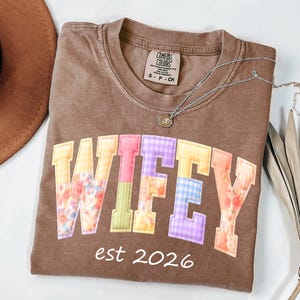 May include: A brown Comfort Colors t-shirt with the word "WIFEY" in colorful patchwork letters. The text "est 2026" is below the word. A silver necklace with a small pendant is on the shirt.