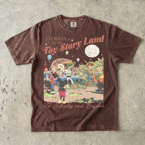 May include: A brown short-sleeve t-shirt featuring a graphic print with the text "FLORIDA Est. 2018 Toy Story Land" and "To Infinity and Beyond". The illustration depicts Mickey Mouse, Woody, Buzz Lightyear, and Slinky Dog under a large moon.