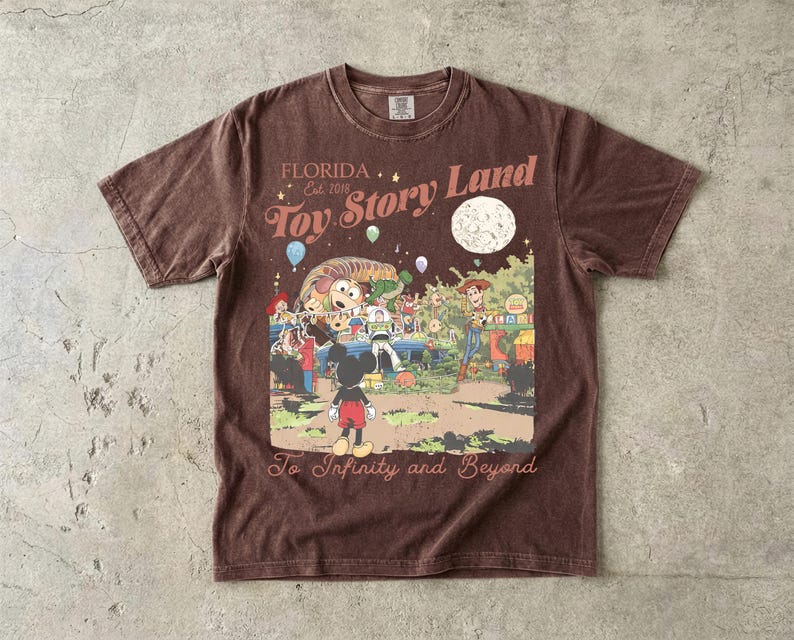 May include: A brown short-sleeve t-shirt featuring a graphic print with the text "FLORIDA Est. 2018 Toy Story Land" and "To Infinity and Beyond". The illustration depicts Mickey Mouse, Woody, Buzz Lightyear, and Slinky Dog under a large moon.