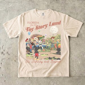 May include: A beige short-sleeve t-shirt featuring a graphic print with the text "FLORIDA Est. 2018 Toy Story Land" and "To Infinity and Beyond". The illustration depicts Mickey Mouse, Woody, Buzz Lightyear, and Slinky Dog in a park setting.