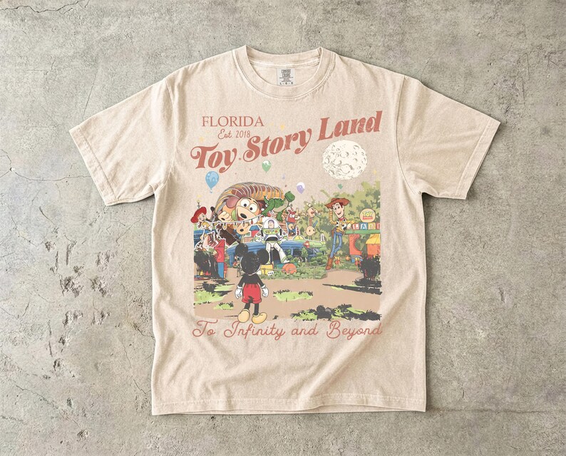 May include: A beige short-sleeve t-shirt featuring a graphic print with the text "FLORIDA Est. 2018 Toy Story Land" and "To Infinity and Beyond". The illustration depicts Mickey Mouse, Woody, Buzz Lightyear, and Slinky Dog in a park setting.