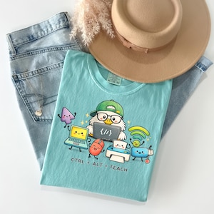 May include: A light blue t-shirt featuring a graphic of an owl wearing a green cap and glasses, surrounded by animated computer accessories including a laptop, mouse, printer, and Wi-Fi symbol, with the text CTRL + ALT + TEACH printed below.