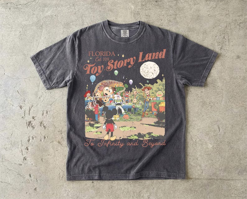 May include: A charcoal gray short-sleeve t-shirt featuring a graphic print with the text Florida Est. 2018 Toy Story Land and To Infinity and Beyond. The illustration depicts characters including Mickey Mouse, Woody, Buzz Lightyear, and Slinky Dog.