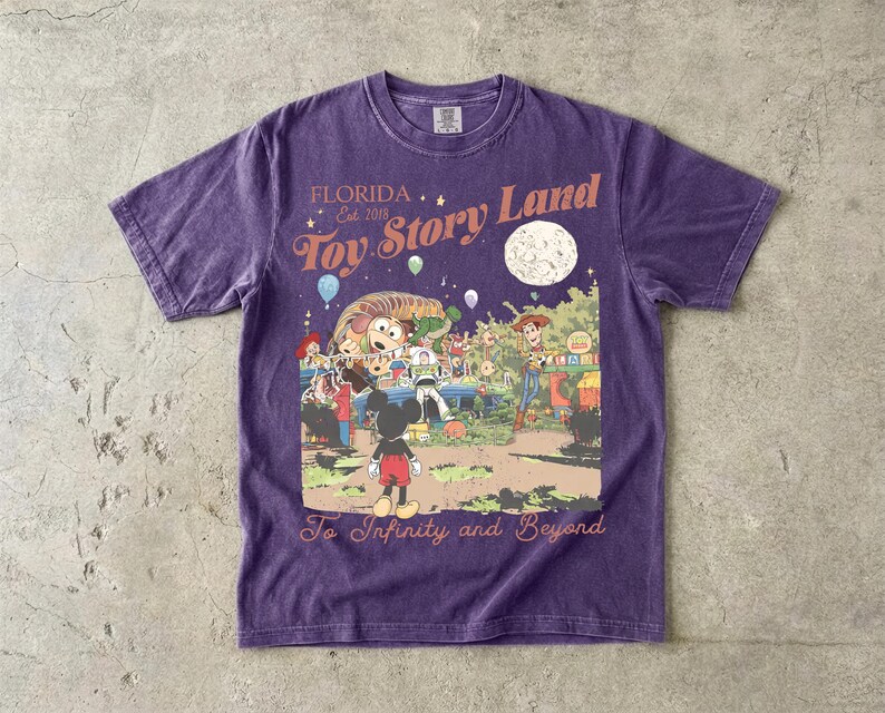 May include: A purple short-sleeve t-shirt featuring a graphic print with the text Florida Est 2018 Toy Story Land and To Infinity and Beyond. The illustration depicts Mickey Mouse, Woody, Buzz Lightyear, and other characters in a park setting.