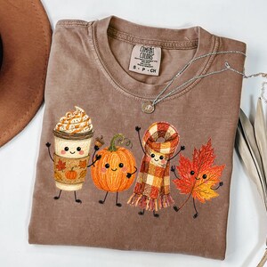 May include: A brown short-sleeve t-shirt featuring a graphic print of a coffee cup, a pumpkin, a plaid scarf, and a maple leaf, each with a smiling face and stick arms and legs. The shirt has a Comfort Colors brand label at the inner neckline.