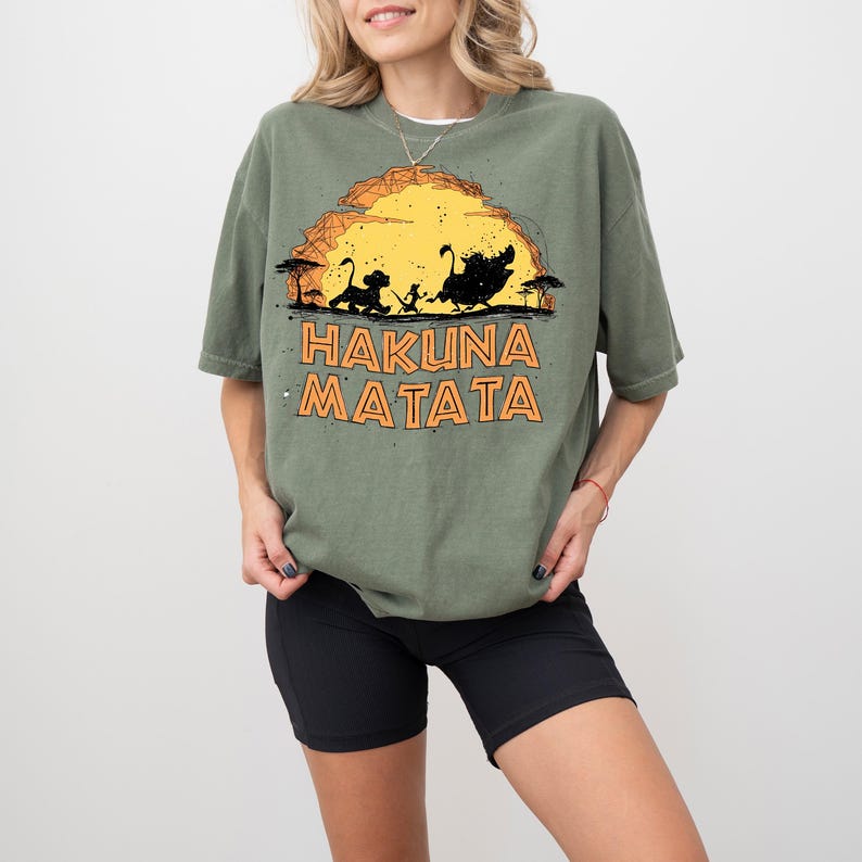May include: An olive green t-shirt featuring a graphic of a yellow sun setting behind silhouettes of a lion cub, a meerkat, and a warthog. Below the graphic, the text HAKUNA MATATA is printed in bold, orange, outlined capital letters.