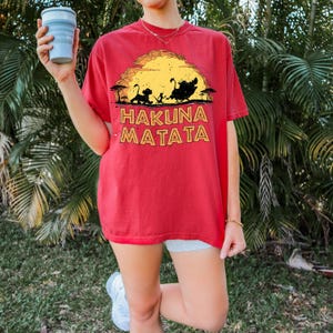 May include: A red short-sleeve t-shirt featuring a graphic of a lion, a meerkat, and a warthog silhouetted against a yellow sun. The text "HAKUNA MATATA" is printed in large yellow letters below the graphic. The shirt has a crew neckline.
