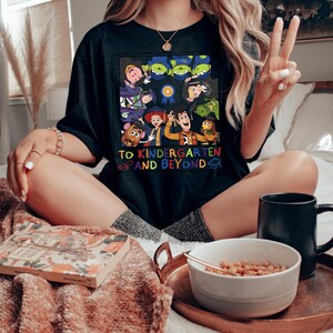 May include: A black t-shirt featuring a colorful graphic of various animated characters wearing graduation caps. The design includes a blue ribbon icon and the text "TO KINDERGARTEN AND BEYOND" in multi-colored letters, accompanied by a small diploma and cap icon.