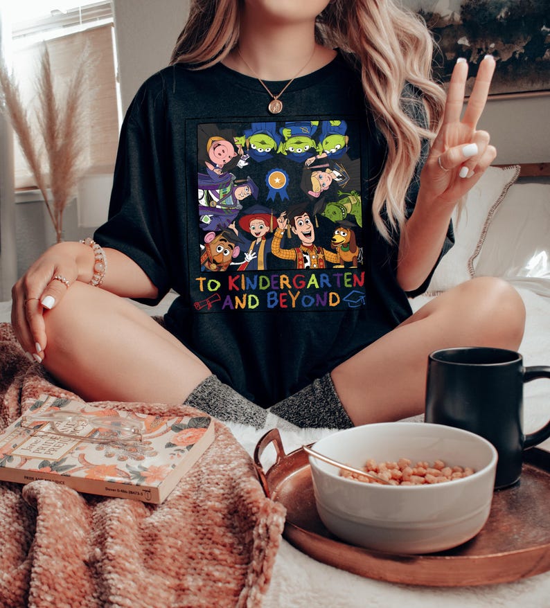 May include: A black t-shirt featuring a colorful graphic of various animated characters wearing graduation caps. The design includes a blue ribbon icon and the text "TO KINDERGARTEN AND BEYOND" in multi-colored letters, accompanied by a small diploma and cap icon.