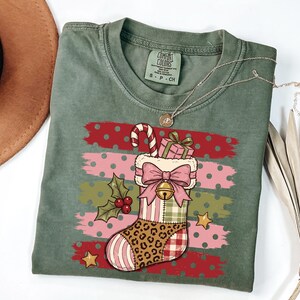 May include: An olive green t-shirt featuring a graphic of a Christmas stocking with a leopard print toe, plaid heel, and pink bow. The stocking contains a candy cane and a gift box, accented by holly berries, stars, and horizontal stripes.