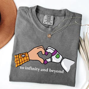 Könnte beinhalten: Ein anthrazitfarbenes T-Shirt mit Rundhalsausschnitt und einer Grafik von zwei Händen, die ein Herz bilden. Eine Hand trägt einen gelb karierten Ärmel, die andere ist eine weiße Roboterhand mit lila und grünen Akzenten. Der Text lautet to infinity and beyond.