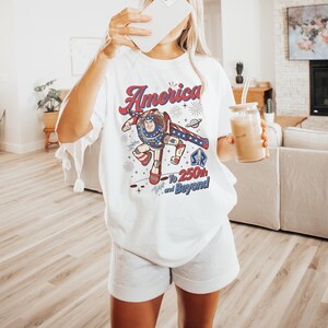 May include: A white t-shirt featuring a graphic of a space ranger character in a red, white, and blue suit. The text reads America To 250th and Beyond, accompanied by star and firework illustrations. The design includes a small SR logo emblem.