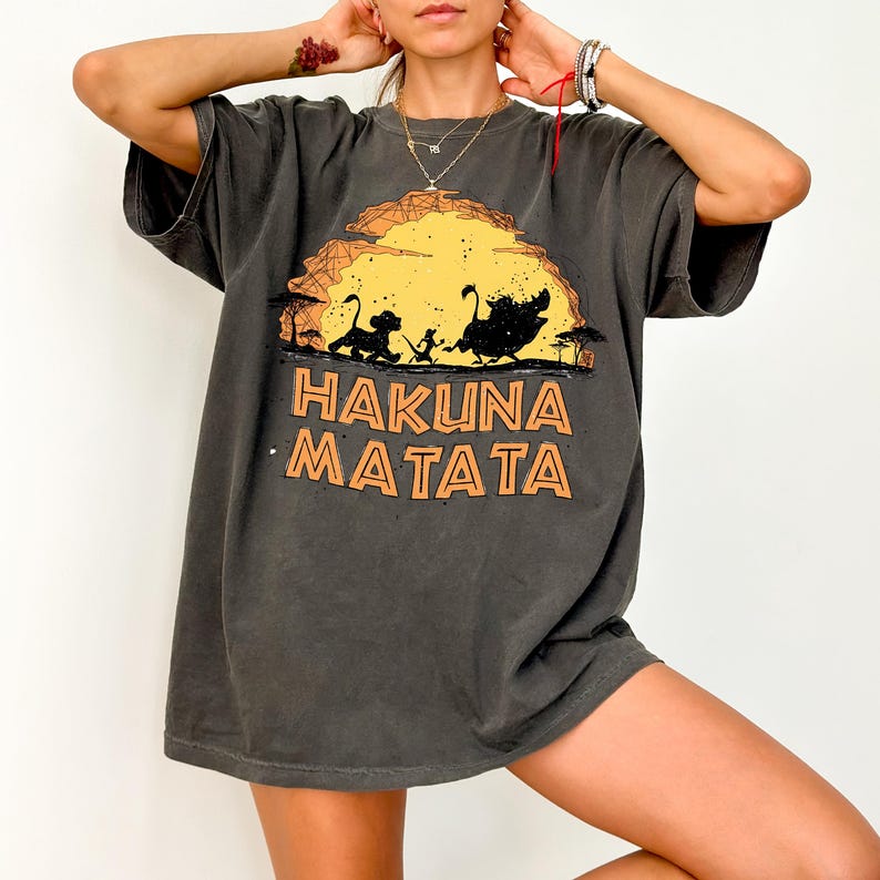 May include: A charcoal gray short-sleeve t-shirt featuring a graphic print of three animal silhouettes walking against a large yellow sun. Below the graphic, the text HAKUNA MATATA is printed in a bold, orange, outlined sans-serif font.