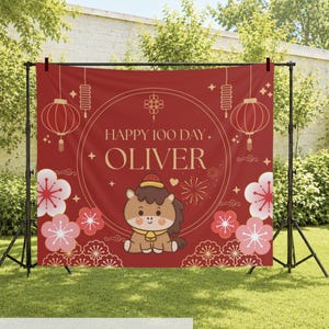 May include: A red rectangular backdrop featuring the text HAPPY 100 DAY OLIVER in gold lettering. The design includes a central cartoon horse, gold lanterns, and pink floral patterns. The backdrop is mounted on a black metal stand frame.