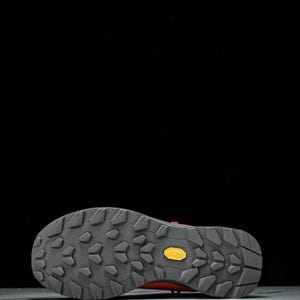 May include: Close-up of the sole of a hiking boot. The sole is grey with a textured pattern and a yellow "Vibram" logo. The boot's upper is a reddish-orange colour, and the background is black.