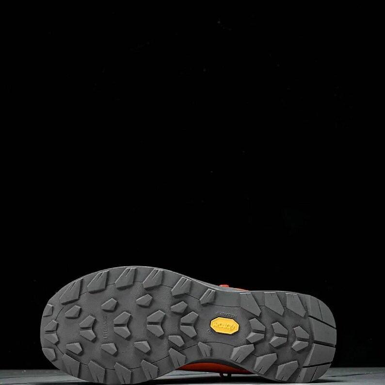 May include: Close-up of the sole of a hiking boot. The sole is grey with a textured pattern and a yellow "Vibram" logo. The boot's upper is a reddish-orange colour, and the background is black.