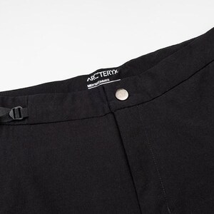 May include: Close-up of black pants with a button closure and a small belt. The pants have a zipper pocket and a brand label that reads "Arc'teryx". The pants are made of a durable fabric.