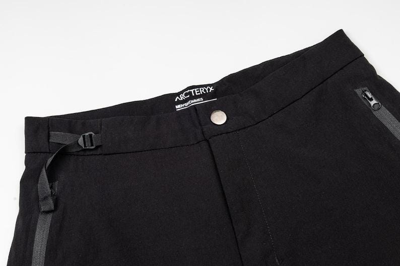 May include: Close-up of black pants with a button closure and a small belt. The pants have a zipper pocket and a brand label that reads "Arc'teryx". The pants are made of a durable fabric.