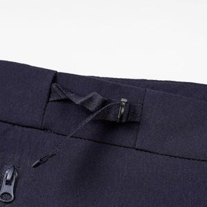 May include: Close-up of navy blue fabric, possibly pants or shorts, featuring a drawstring waistband and a zippered pocket. The fabric has a subtle texture, and the zipper pull is visible.