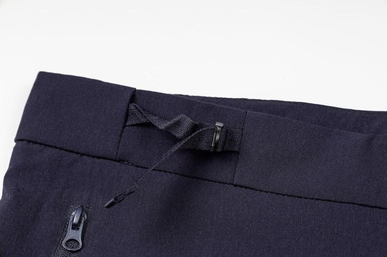 May include: Close-up of navy blue fabric, possibly pants or shorts, featuring a drawstring waistband and a zippered pocket. The fabric has a subtle texture, and the zipper pull is visible.